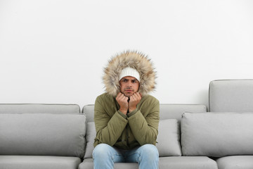 Young man in warm clothes freezing on sofa against white background