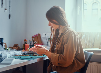Attractive girl is making necklace from beads and watching instructions on computer.