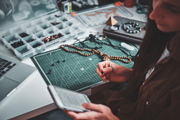 Young woman is chatting by mobile phone while sitting next to table with her jewellery hobby.