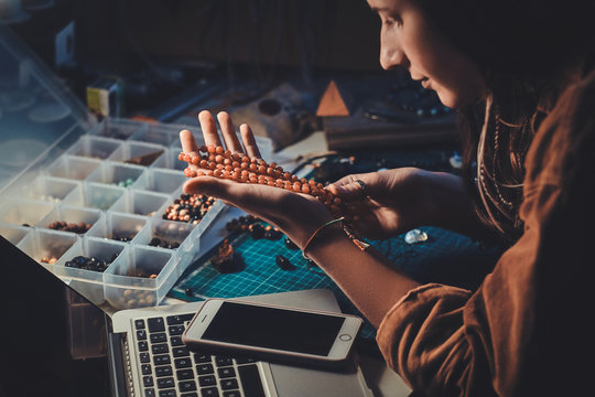 Talented girl is holding handmade beaded necklace in hand while sitting at her workshop.