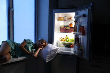 Woman sleeping on window sill near open refrigerator in kitchen at night