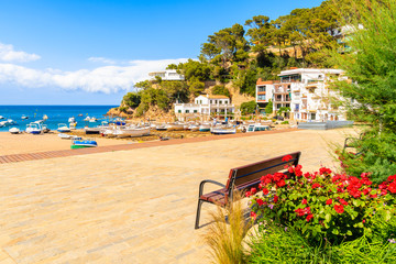 Flowers and bench on coastal promenade and view of fishing boats on beach in beautiful Sa Riera village, Costa Brava, Catalonia, Spain