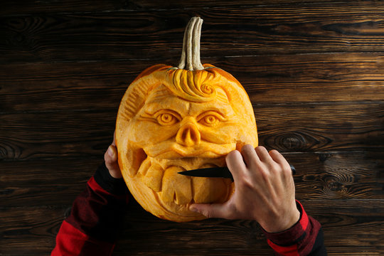 Top view shot of male hands carving creative human face shaped jack-o-lantern with big croocked teeth. Funny halloween pumpkin being carved by unrecognizable man. Close up, copy space, wood background