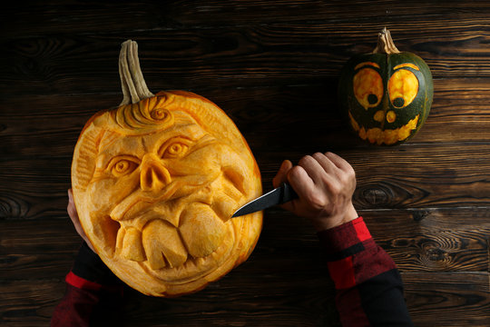 Top view shot of male hands carving creative human face shaped jack-o-lantern with big croocked teeth. Funny halloween pumpkin being carved by unrecognizable man. Close up, copy space, wood background