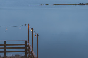 Twilight, gray color sky at the lakescape, electric lamps as garland and wooden terrace, pier. romantic atmosphere of evening. Night cafe terrace. Retro garland for home decor.