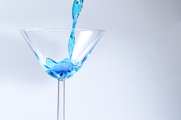 Cocktail with blue liquid in glass. Glass with blue water pouring with liquid with splashes and drops. Martini glass filling with alcohol with splashes on white background. Refreshing drink concept.