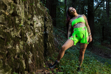 Naklejka premium Asian tattooed woman in neon rave clothing poses on an abandoned rock wall in the forest