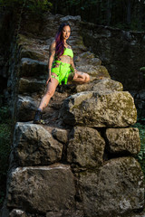 Asian tattooed woman in neon rave clothing poses on an abandoned rock wall in the forest