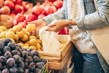 Female hands puts fruits and vegetables in cotton produce bag at food market. Reusable eco bag for shopping. Sustainable lifestyle. Eco friendly concept.