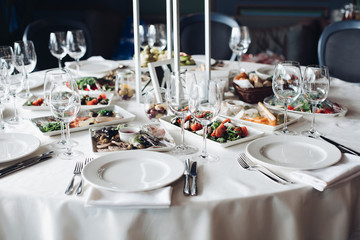 View over wedding table covered with white tablecloth with empty glasses, plates and utensils. Snacks are served on the table.
