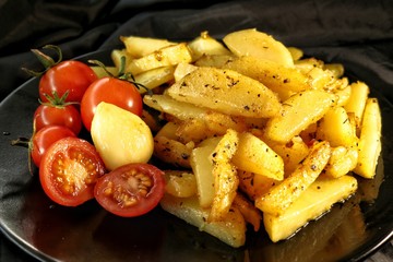 fried potatoes with crust, spices, garlic and fresh cherry tomatoes on a black plate