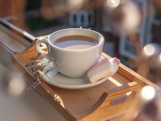 White cup of coffee is situated on the wooden tray at the windowsill in the rays of morning sun,...