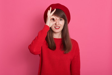 Close up portrait of young woman looking through OK sign, adorable female dresses red clothing and beret, attractive girl covering her eye with ok gesture, posing isolated over pink background.