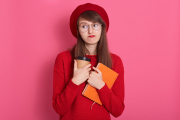Photo of thoughtful Caucasian teenage girl with dark staraight hair, wearing red beret and sweater, isolated over pink background, looks aside, copy space for promotion text or advertisment.
