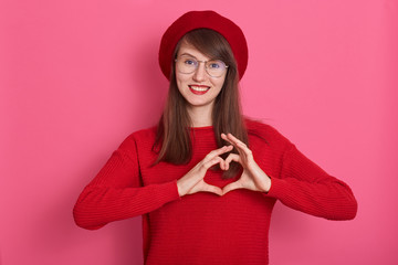 Close up portrait of beautiful young woman posing isolated over rosy background, female doing heart shape with her hands, showing love gesture to her boyfriend or hausband. Human feelings concept.