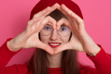 Close up portrait of beautiful woman with dark staright hair,wearing red headdress, shirt, spectacles, making heart shapes with her hands in front of her face, isplated over rosy background in studio.