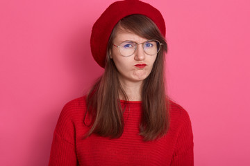 Portrait of sullen angry offended girl frowns face, pouts lips, being irritated,posing isolated over pink background, discontent young woman with gloomy expression after quarrel, dresses red clothing