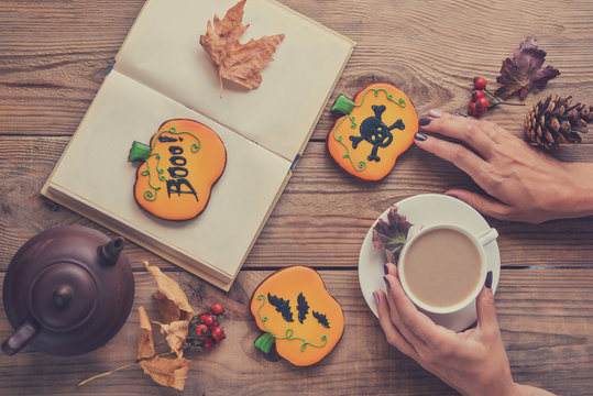 Halloween cookies and cup of coffee on the table. Women's hands, a cup of coffee, a book, Halloween postcard, toned image