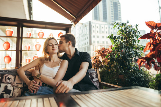 Cute loving couple sitting in cozy restaurant and terrace on sunset background and waiting for order. Young man hugs and kisses a girl in a cozy cafe, she smiles.