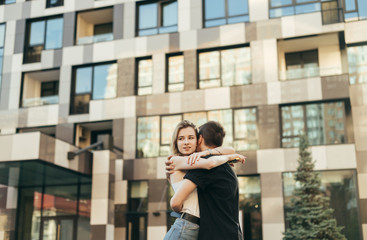 Beautiful young couple hugging on the street on the background of a modern building, the girl looks away. Guy hugged the girl on a walk on a warm summer day, she is happy. Love story