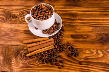 White cup filled with coffee beans, star anise and cinnamon sticks on wooden table