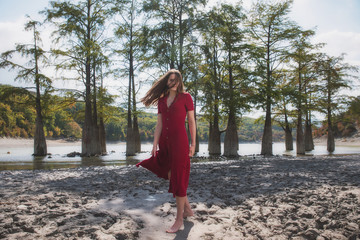 an expressive portrait of a young brown-haired woman in a red dress on the background of a morning lake with cypresses, a girl with a slender figure posing against nature