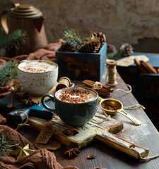 christmas still life with two cups of hot creamy drink (cocoa, chocolate, coffee) with snowflake cocoa ornament on top stands on wooden boards on rustic table with fir tree, spices