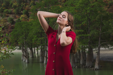a beautiful, young brown-haired woman in a red dress on the background of a morning lake with cypresses, a girl with a slender figure posing against nature