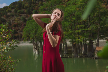 a beautiful, young brown-haired woman in a red dress on the background of a morning lake with cypresses, a girl with a slender figure posing against nature