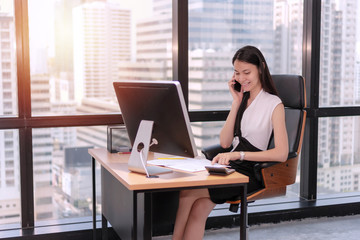 Professional young businesswoman sitting in the office room with desktop monitor and document while use smart phone talking about job on city background. Business accomplishment concept.