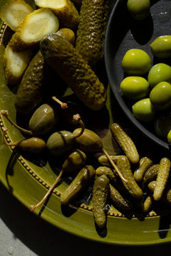 A selection of pickled gherkins, caper berries and green olives on an olive green plate.