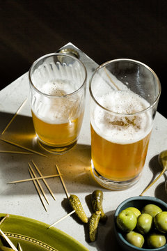 Beer and snacks. A selection of pickled gherkins, caper berries and green olives accompany two glasses of beer on a concrete background.
