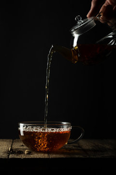 A refreshing cup of chai tea being poured from a glass teapot into a glass teacup. The surface is rustic timber.