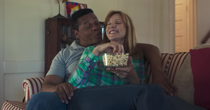 Happy Older Couple In Their 50s And 60s Eating Popcorn While Sitting Back On The Couch Together Watching A Movie. Wife Feeding Her Husband A Snack While They Cuddle On The Sofa. Slow Motion 4k