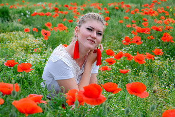 Blonde young woman in red skirt and white shirt, red earrings is in the middle of a poppy field.