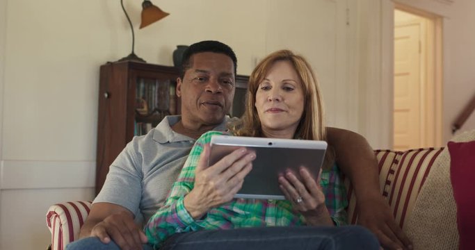 Happy Older Couple Streaming Movie Or Show Online With Tablet Computer While Sitting On Couch. African American And Caucasian Husband And Wife Watching News On Mobile Device. Slow Motion