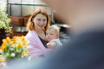 Close up of mother carrying her baby daughter in a pink sling wrap while shopping for flowers for their garden in a local greenhouse