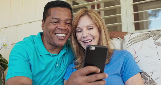 Close up of happy older couple sitting on porch and using smartphone together. African American and Caucasian husband and wife looking at cell phone on sunny day outside home. Slow motion 4k handheld