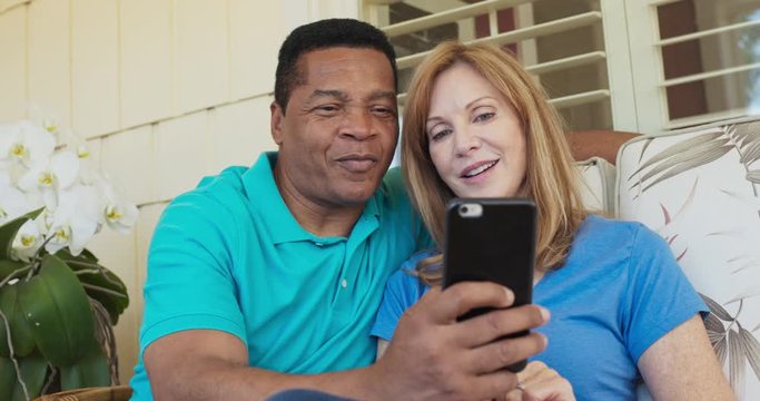 Smiling older couple sitting on porch and using cell phone and laughing together. African American and Caucasian husband and wife looking at smartphone on sunny day outside home. Slow motion 4k