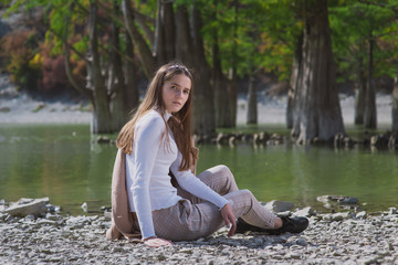 a beautiful, young brown-haired woman in a beige coat and a white turtleneck poses against a lake with cypresses in dark ankle boots