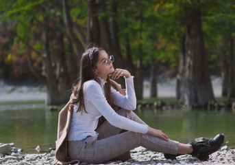 a beautiful, young brown-haired woman in a beige coat and a white turtleneck poses against a lake with cypresses in dark ankle boots