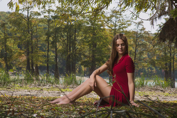 beautiful, young brown-haired woman in a red dress on the background of the morning lake with cypresses and sun rays in the fog