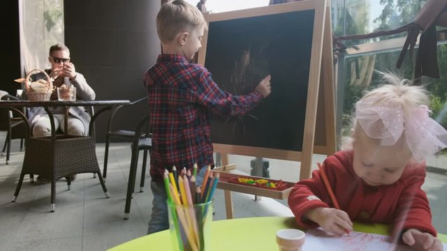 A boy and a girl are playing at the table on the veranda. Sister draws with pencils, and brother draws with chalk on the blackboard.