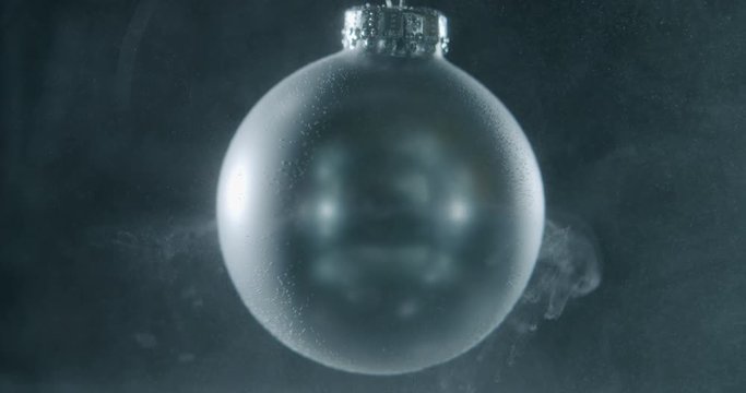 Silver Christmas bauble slowly spinning in a dark environment. Small particles in the orbit of the bauble. Other particles falling onto the bauble.