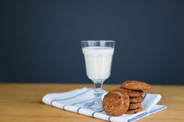 round brown wholemeal oatmeal cookies stack with raisins and white milk in glass cup on striped textile cloth on wooden table, close up side view of vertical still life stock photo image
