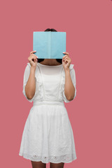 young woman in dress covering face while reading blue book isolated on pink