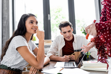 multiethnic students smiling and looking at laptop in apartment