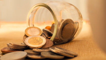 A coin jar overturned on the table with euro coins. This image can be used to represent different...