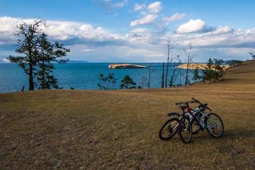 Obraz premium View of the island on Lake Baikal across the steppe. In the field they stand embracing two bicycles. At the edge of the coast trees grow, on the horizon of the mountain. Clouds in the sky.