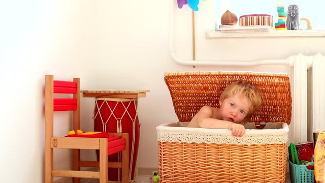 Beautiful blonde caucasian funny little male child hiding in handmade straw basket playing in kids room, smiling and looking at camera magnificent blue eyes. Happy family childhood. Modern interior.